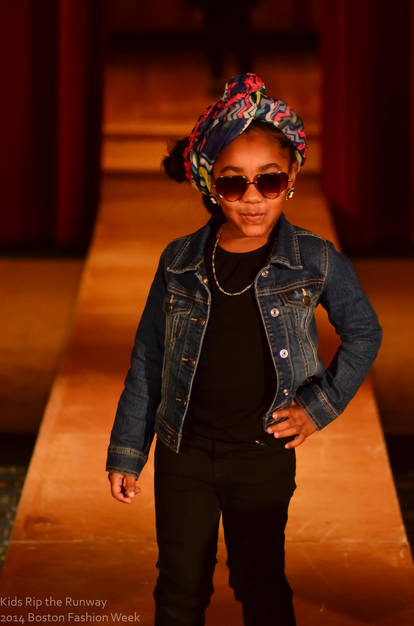 A young model posing confidently on the runway in sunglasses and a denim jacket.